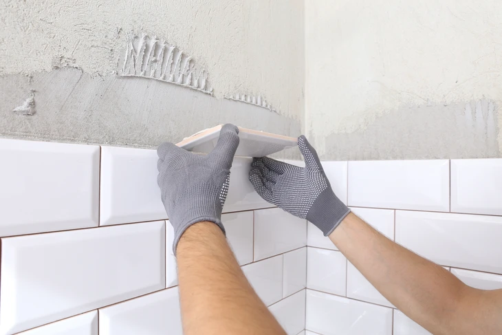 Un plombier en salopette installe ou répare une robinetterie de douche dans une salle de bain carrelée blanche
